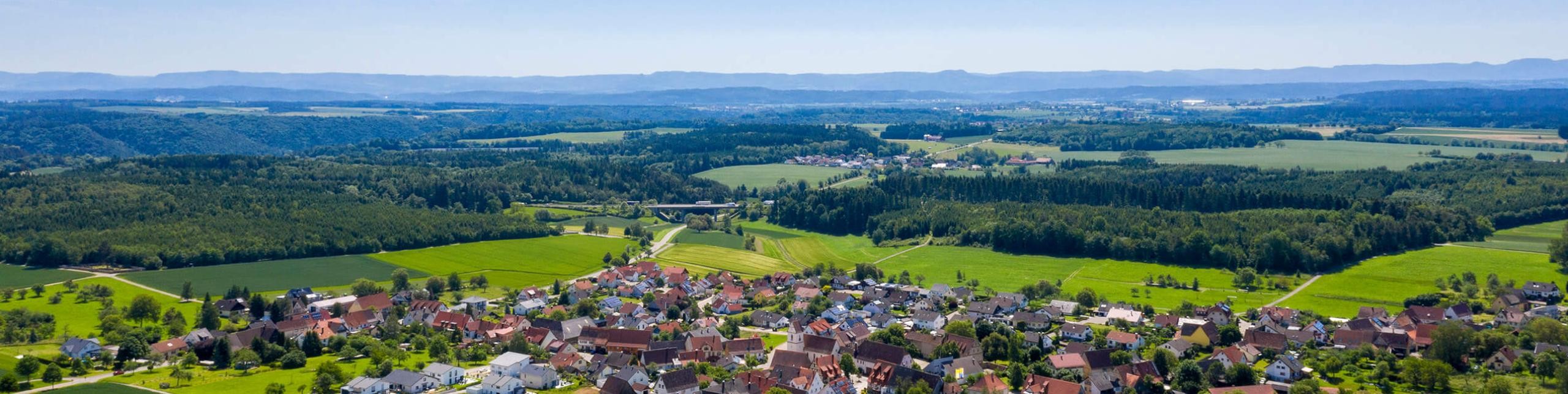 Luftaufnahme einer Ortschaft in der ländlichen Umgebung, umgeben von grünen Feldern und sanften Hügeln.
