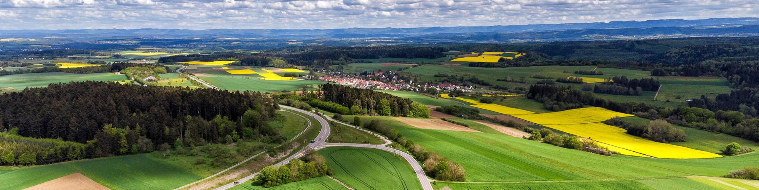 Luftaufnahme einer kleinen Ortschaft in der ländlichen Umgebung, umgeben von grünen Feldern und sanften Hügeln.