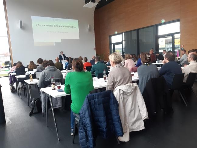 Das Bild zeigt den Veranstaltungssaal aus der hinteren Perspektive. Die Teilnehmer sitzen mit dem Rücken zur Kamera und blicken nach vorne, wo Landrat Andreas Junt seine Rede hält. Über ihm ist eine Präsentationsfolie zu sehen.