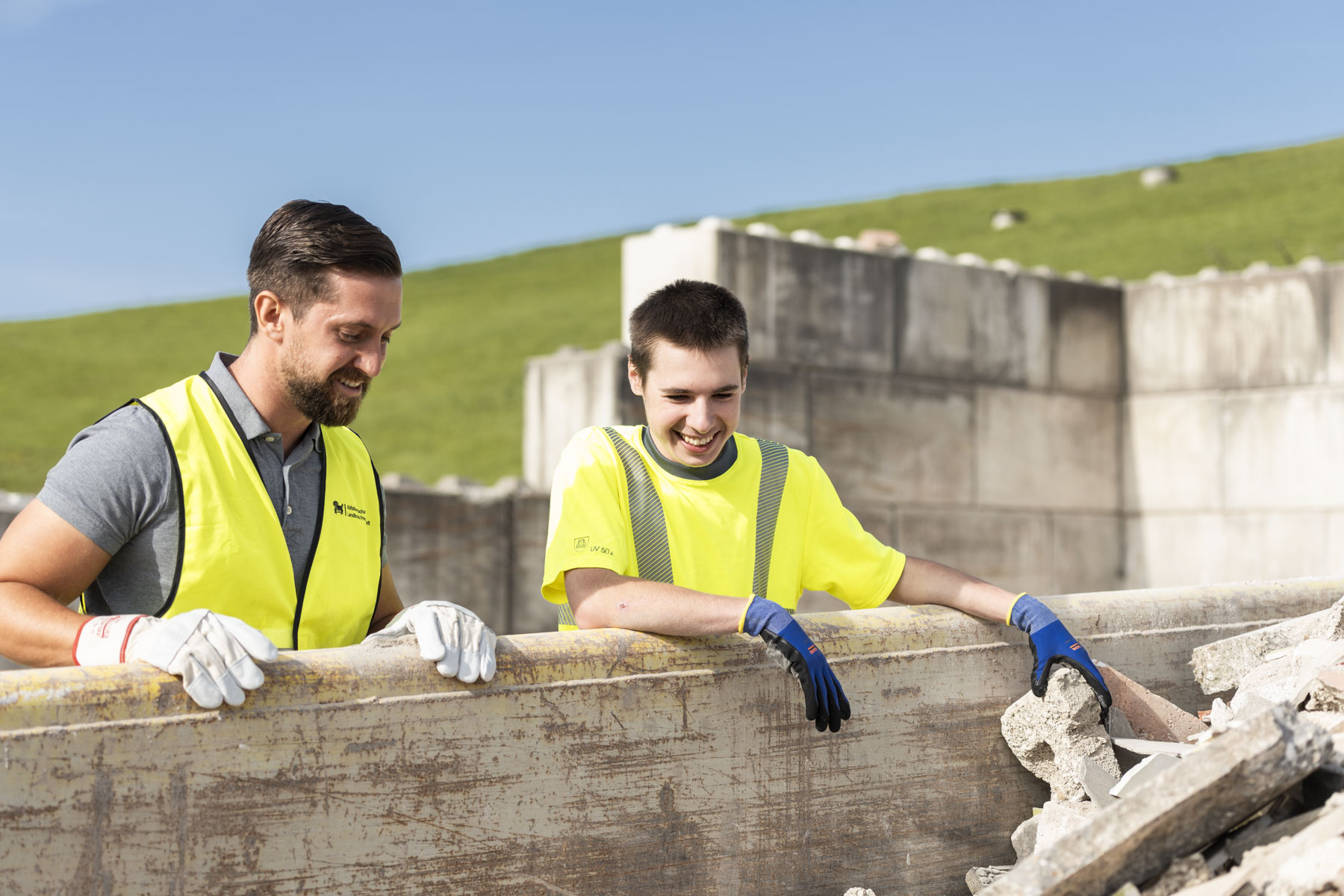 Zwei Umwelttechnologen mit gelber Warnweste stehen an einem Container und schauen in diesen hinein.